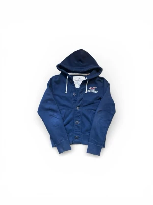 Marinblå hoodie från Hollister - Snygg marinblå hoodie från Hollister med knappar framtill och vit dragsko i huvan. Broderad logga och text på bröstet. Tillverkad i mjuk bomullsblandning och har kängurufickor. Perfekt för en chill och avslappnad stil.