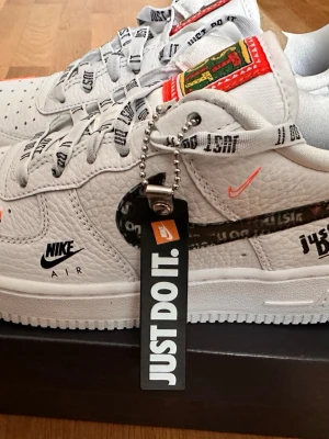 Nike Air Force 1 Just Do It sneakers - Snygga vita Nike Air Force 1 sneakers med 'Just Do It' detaljer på snören, sidor och tag. Skorna har svart swoosh med text, orange och svarta loggor samt färgglad patch på plösen. Tillverkade i läder med klassisk låg siluett och perforerad tå.  Modellen släpptes juni 2018 för att fira 30 års jubileum med Nike’s ikoniska slogan ”Just Do It”. Helt nya oanvända, orginalkartonnocj original tillbehör.