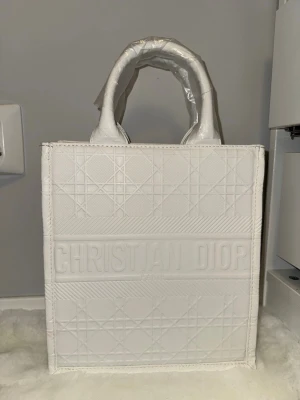 Vit handväska - Säljer en snygg vit handväska med klassiskt geometriskt mönster och tydlig logga framtill. Väskan har två robusta handtag och en boxig form, perfekt för dig som vill ha en trendig och stilren accessoar. 