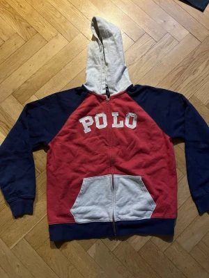 Röd och blå hoodie från Polo Ralph Lauren - Snygg hoodie från Polo Ralph Lauren med röd kropp, marinblå ärmar och vit huva. Stor POLO-text i vitt på bröstet och stor ficka framtill. Dragkedja hela vägen och kontrastfärgad ficka och huva. Perfekt för en avslappnad streetwear-look.