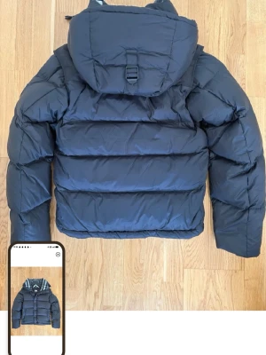 Svart pufferjacka från Burberry - Snygg svart pufferjacka från Burberry med stor huva och tydlig logotyp på ärmen. Jackan har breda kanaler, dragkedja och tryckknappar samt en urban look. Perfekt för kalla dagar när du vill hålla stilen och värmen.