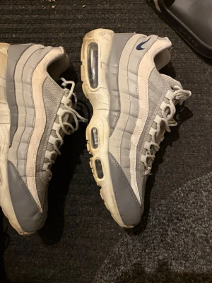 Nike Air Max 95 grå/vit sneakers - Nike Air Max 95 sneakers i grått och vitt med ikoniska vågformade paneler och synlig Air-dämpning i sulan. Ovandel i mesh och läder, klassisk snörning och diskret Swoosh-logga på sidan. Perfekta för dig som gillar streetwear och sportig stil. Köp gärna med instabox så skickas det samma dag om köpet sker innan 16!
