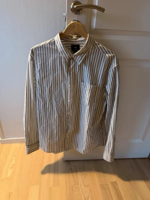 Randig skjorta från H&M i bomull - Säljer en klassisk långärmad skjorta från H&M med vertikala ränder i beige och vit. Skjortan har knappar framtill, bröstficka och en avslappnad passform. Perfekt för dig som gillar stilrena och enkla plagg.