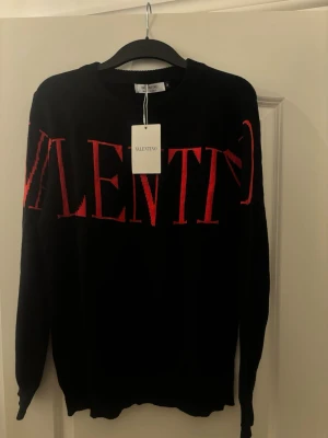Svart stickad tröja från Valentino - Snygg svart stickad tröja från Valentino med stort rött VALENTINO-tryck över bröstet. Tröjan har rund halsringning och långa ärmar. Perfekt för dig som gillar exklusiva och trendiga plagg med tydlig logga.