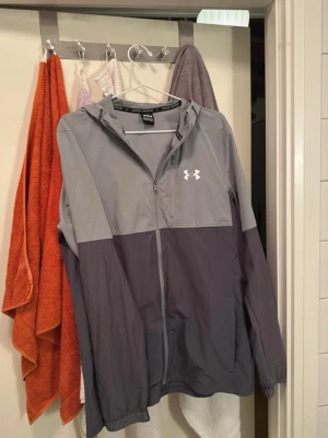 Grå vindjacka från Under Armour - Snygg tvåfärgad vindjacka från Under Armour i ljusgrått och mörkgrått. Jackan har huva, dragkedja framtill och logga på bröstet. Tillverkad i lätt och tåligt syntetmaterial, perfekt för blåsiga dagar.
