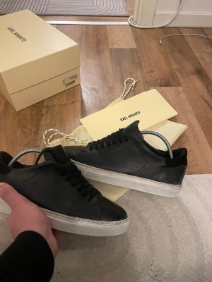 Svarta Axel Arigato sneakers i läder - Snygga svarta sneakers från Axel Arigato i slimmad design. Tillverkade i slätt läder med svart ovandel och vit sula. Klassisk låg modell med svarta skosnören och diskret logga på sidan. Perfekta för en clean och stilren look. Storlek 40 passar 41