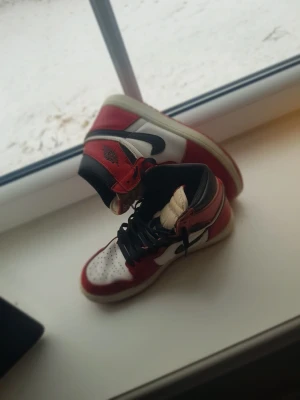 Air Jordan 1 Chicago röda sneakers - Säljer ett par Air Jordan 1 Chicago sneakers från Nike. Skorna har klassisk high-top design med röd, vit och svart färgkombination, svart swoosh och perforerad tå. Materialet är läder och textil, med ikonisk logga på sidan. Perfekt för dig som gillar streetstyle.