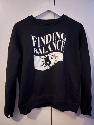 Svart sweatshirt med tryck - Cool svart sweatshirt med vitt tryck på bröstet där det står 'FINDING BALANCE' och ett yin-yang-motiv med solstrålar. Tröjan har rund halsringning, långa ärmar och ribbade muddar. Perfekt för en avslappnad streetwear-look.