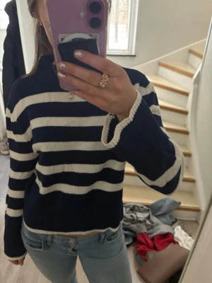 Marinblå och vit randig stickad tröja - Säljer en stickad tröja med breda marinblå och vita ränder. Tröjan har en rak passform, ribbad halsringning och muddar vid ärmslut och nederkant. Perfekt till jeans för en avslappnad look. Materialet känns mjukt och bekvämt.