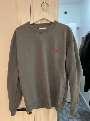 Ami Paris Crewneck Sweatshirt - Säljer en stilren grå/blå sweatshirt från Ami Paris med det ikoniska röda hjärtat och bokstaven A broderat på bröstet. Tröjan har rund halsringning, ribbade muddar och är tillverkad i bomull. 