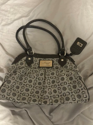 Grå handväska  - Snygg handväska  i grått tyg med svart GG-mönster och blommor. Väskan har svarta detaljer i konstläder, dubbla handtag och en bricka som har en spegel. Praktisk dragkedja på baksidan. Vet ej vad det är för märke, fick den av familjemedlem. Aldrig använts, säljs pågrund av att den ej används