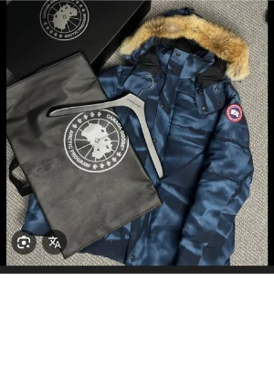  Canada Goose whyndham - Säljer en blå parkas från Canada Goose med snyggt mönster och stor pälsluva. Jackan har flera fickor, knappar och dragkedja framtill samt den klassiska loggan på ärmen. Perfekt för kalla dagar och riktigt stilren.