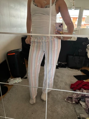 Randiga linnebyxor i pastellfärger - Säljer ett par luftiga linnebyxor med raka ben. Byxorna har ett vitt basmaterial med tunna ränder i pastellrosa, blått, gult och lila. Perfekta för en avslappnad och trendig look. Materialet känns lätt och svalt, perfekt för varmare dagar. Står storlek 146 men passar xs