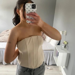 Beige axelbandslös korsett - Säljer en stilren beige korsett med markerade sömmar och formande passform. Axelbandslös design som ger en snygg siluett och passar perfekt till jeans eller kjol. Materialet är slätt och mjukt, vilket gör den bekväm att bära.