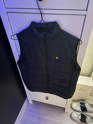 Svart dunväst från Lyle & Scott - Snygg svart dunväst från Lyle & Scott med gul broderad logga på bröstet. Västen har hög krage, dragkedja framtill och quiltad design. Perfekt att slänga över en hoodie för extra värme. Klassisk och stilren look.
