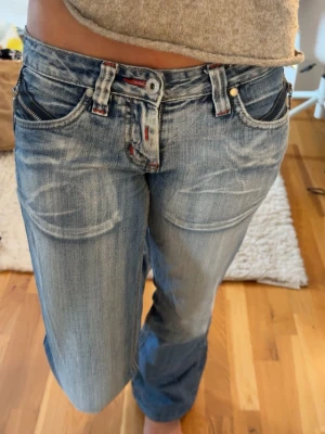 Blåa loose fit jeans från - Säljer ett par blåa loose fit jeans  med slitningar och ljusa tvättade detaljer. Jeansen har låg midja, raka ben och röda sömmar vid fickorna. Klassisk femficksmodell med dragkedja och knappgylf.