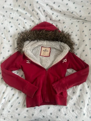 Vintage röd hoodie med fuskpäls från Hollister - Säljer en vintage röd hoodie från Hollister med dragkedja, mysigt vitt teddyfoder och huva med brun fuskpäls. Hollister-logga broderad på bröstet. Perfekt för dig som vill ha en varm och snygg hoodie med skön passform och coola detaljer. Vintage storlek L passar mer som S måtten finns. 