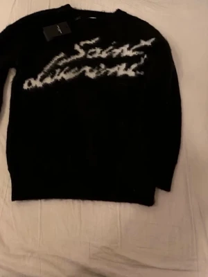 Svart Saint Laurent mohairtröja - Svart långärmad tröja från Saint Laurent i mjuk mohairblandning. Tröjan har vit, suddig logotyptext framtill och ribbade muddar vid ärmslut och nederkant. Oversized och trendig passform, perfekt för en cool streetstyle-look.