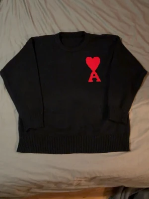 Svart stickad tröja Ami Paris - Svart stickad tröja från Ami Paris med röd hjärtlogga och bokstaven A på bröstet. Tröjan har rund halsringning, ribbade muddar och nederkant. Perfekt för dig som gillar stilrena plagg med en twist.