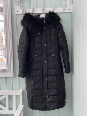 Rockandblue Lång Dunjacka Svart Strl 38 - Snygg lång svart dunjacka från Rockandblue med quiltade detaljer och huva med avtagbar svart pälskant. Dragkedja och knappar framtill samt flera fickor med dragkedja. Varm och funktionell med en stilren, smickrande silhuett – perfekt för kalla vinterdagar. Mycket fint skick.