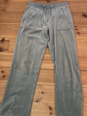 Gröna/turkos velourbyxor Juicy Couture - Mjukisbyxor i grön/turkos velour från Juicy Couture med bred resår i midjan och dragsko. Stora fickor fram och bak, bakfickorna har knappdetaljer. Perfekta för en chill och avslappnad stil.