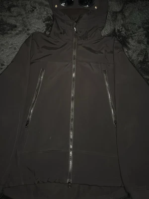 Svart vindjacka med huva och glasögon - Cool svart vindjacka från C.P. Company med huva som har inbyggda glasögon och dragkedja framtill. Jackan har två snedställda fickor med dragkedja och detaljer i metall. Perfekt för dig som vill sticka ut och gillar streetwear.