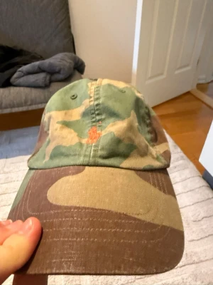 Klassisk camo keps från Polo Ralph Lauren - Säljer en camouflagemönstrad keps från Polo Ralph Lauren. Kepsen har klassisk böjd skärm och justerbar rem bak. Tillverkad i bomull med gröna, bruna och beige toner samt orange detaljer på insidan. Perfekt för dig som gillar streetwear och tidlös stil.