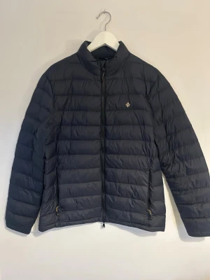 Marinblå dunjacka med dragkedja från Morris  - Snygg marinblå dunjacka från Morris med en diskret logga på bröstet. Jackan har hög krage, två sidofickor med dragkedja och hel dragkedja framtill. Perfekt för kalla vinterdagar och riktigt lätt att bära. Passar perfekt när man vill ha en lite mer diskretare jacka 