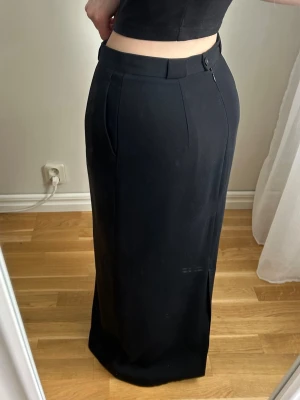 Svart långkjol med slits - Stilren svart långkjol med hög midja och diskret slits på sidan. Kjolen har bälteshällor och stängs med dragkedja i sidan. Perfekt för en clean och klassisk look.