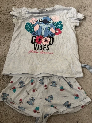 Stitch pyjamas set med tryck - Grå pyjamas med kortärmad t-shirt och shorts, båda med Stitch-motiv från Disney. T-shirten har färgglada blommor, gröna blad och texten 'Good Vibes Aloha Hawaii'. Shortsen har matchande Stitch- och blomtryck. Perfekt för mysiga kvällar.