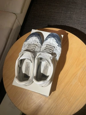 Maison Margiela gats  - Unika vita sneakers från Maison Margiela med blå färgstänk över hela skon. Klassisk låg modell med vita snören och detaljerade sömmar. Tillverkade i läder för en lyxig känsla och cool look. Perfekta för dig som vill sticka ut med din stil.