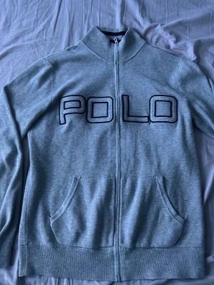 Grå ziptröja från Polo Ralph Lauren - Snygg grå ziptröja från Polo Ralph Lauren med stor POLO-text på bröstet. Tröjan har ribbade muddar, två fickor framtill och hög krage med dragkedja. Perfekt för en avslappnad och cool look.