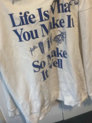 Vit hoodie med blå text och fjäril - Säljer en vit hoodie med blå texten 'Life Is What You Make It So Make It Well' och blå fjärilsmotiv. Hoodien har huva och magficka, samt ribbade muddar. Perfekt för dig som gillar statement-plagg och snygga tryck.