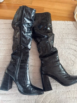 Svarta höga boots med krokoprägling - Snygga svarta stövlar med högt skaft och chunky klack. Stövlarna har ett glansigt krokodilpräglat mönster och dragkedja på insidan. Perfekta för att lyfta din outfit med en edgy vibe. Materialet är konstläder med croco-effekt.