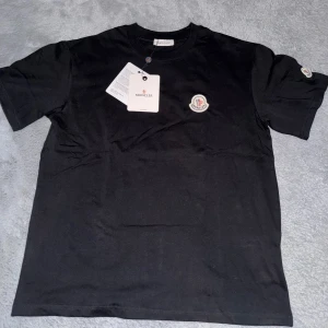 Svart Moncler t-shirt med logga - Svart t-shirt från Moncler i mjuk bomull med klassisk rund hals. Ikonisk Moncler-logga broderad på bröstet och även på ärmen. Enkel och stilren design som passar perfekt till jeans eller shorts.