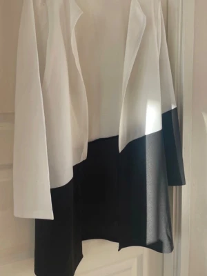 Svartvit kavaj i rak modell - Snygg svartvit kavaj med öppen front och rak passform. Kavajen har en cool färgblock-design med vit ovandel och svart nedtill. Långa ärmar och mjukt, följsamt tyg som ger en modern look. Perfekt  att styla till jeans eller kjol för en trendig outfit.