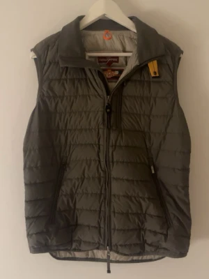 Grå Parajumpers väst - Snygg grå dunväst från Parajumpers med klassisk gul detalj vid dragkedjan. Västen har ståkrage, två sidofickor med dragkedja och quiltad design. Perfekt för lager-på-lager och håller dig varm under kyliga dagar.