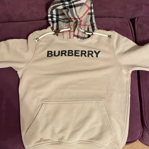 Beige hoodie från Burberry - Säljer en snygg beige hoodie från Burberry med klassiskt rutigt mönster i huvan. Tröjan har en stor ficka framtill och justerbara snören vid huvan. Perfekt för en stilren look. Pris kan diskuteras…