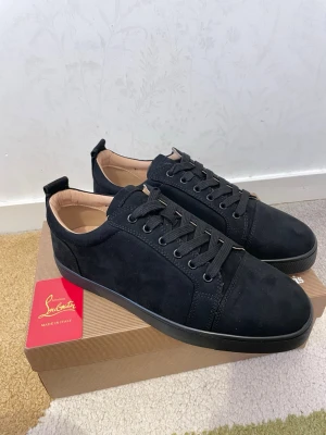 Svarta sneakers från Christian Louboutin - Stilrena svarta sneakers i mocka från Christian Louboutin med klassisk röd sula och svarta skosnören. Låg modell med diskreta detaljer och lyxig känsla. Perfekta för dig som vill ha något exklusivt och tidlöst i garderoben.