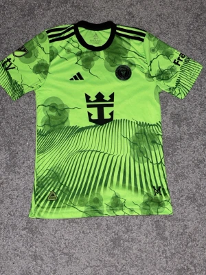 Grön Messi matchtröja Inter Miami - Snygg grön matchtröja från Adidas för Inter Miami med Messi och nummer 10 på ryggen. Tröjan har svarta detaljer, klubbmärke och coolt grafiskt mönster.