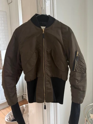 Olivgrön cropped bomberjacka - Snygg olivgrön bomberjacka med croppad passform och svarta ribbade detaljer i midjan och vid kragen. Jackan har guldiga dragkedjor, två stora fickor framtill och en klassisk ficka på ärmen. Bra skick men använd. Storlek M men passar en S också 🖤