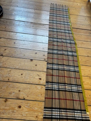 Beige rutig halsduk i klassiskt mönster - Säljer en beige halsduk med det ikoniska rutiga mönstret i svart, vitt och rött. Halsduken är 162 lång, 30 cm bred och har fransar i ändarna. Materialet  är polyester. Knappt använd! Fine skick!