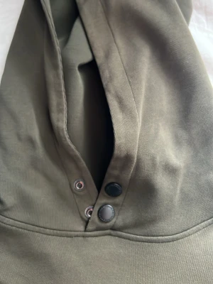 Olivgrön hoodie med knappar - Säljer en olivgrön hoodie med huva och snygga knappar framtill. Hoodien är gjord i mjukt bomullsmaterial och har en stilren design som passar perfekt till jeans eller joggers. Perfekt för dig som gillar en enkel och clean look.