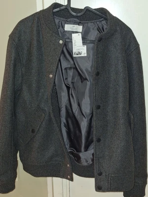 Mörkgrå bomberjacka från H&M - Stilren mörkgrå bomberjacka från H&M med ribbad krage, muddar och nederkant. Jackan har tryckknappar framtill och två snedställda fickor med lock. Materialet är en mjuk ullblandning med slät innerfoder för extra komfort. St 158
