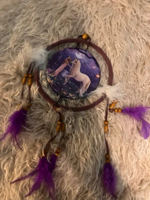 Drömfångare med enhörningsmotiv - En drömfångare med ett motiv av en enhörning och en flicka i mitten, omgiven av lila och vita fjädrar samt träpärlor. Ringen är brun och dekorationerna hänger i läderband. Perfekt för att ge rummet en magisk känsla.