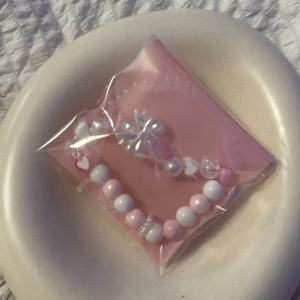 Sött pärlarmband med rosett - Ett gulligt armband med pärlor i vitt, rosa och transparent. Armbandet har en dekorativ rosett, hjärtformade detaljer och skimrande pärlor. Perfekt accessoar för dig som gillar söta och lekfulla smycken. Elastiskt band för enkel påtagning.