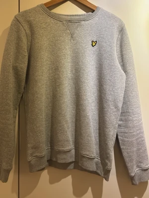Grå sweatshirt från Lyle & Scott - Snygg grå sweatshirt från Lyle & Scott med klassisk rund hals och ribbade muddar. Tröjan har det ikoniska gula örnlogotypen broderad på bröstet. Perfekt för dig som gillar stilrena och bekväma plagg.