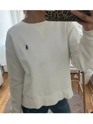 Vit sweatshirt från Ralph Lauren - Klassisk vit sweatshirt från Ralph Lauren med rund halsringning och den klassiska blå loggan broderad på bröstet. Tröjan har en avslappnad passform och ribbade muddar vid ärmslut och nederkant. 