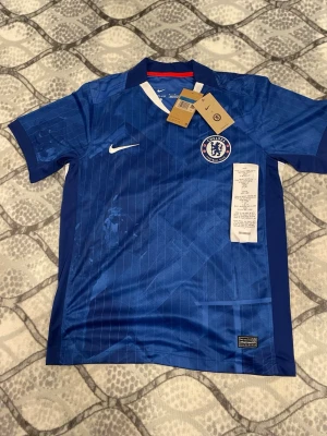 Cole palmer Chelsea fc matchtröja  - Chelsea FC matchtröja från Nike i blått med vita detaljer och tunna vertikala ränder. Tröjan har klubbmärket på bröstet, Nike-logga och klassisk krage med vit V-detalj. Tillverkad i lätt och ventilerande material, perfekt för fotbollsträning.
