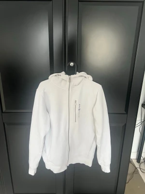 Vit hoodie från Sail Racing - Säljer en vit hoodie från Sail Racing med dragkedja framtill och en bröstficka med zip. Klassisk huva och ribbade muddar vid ärmslut och nederkant. Champion-logga på bröstet. Perfekt för en clean och sportig look. 9/10 skick!
